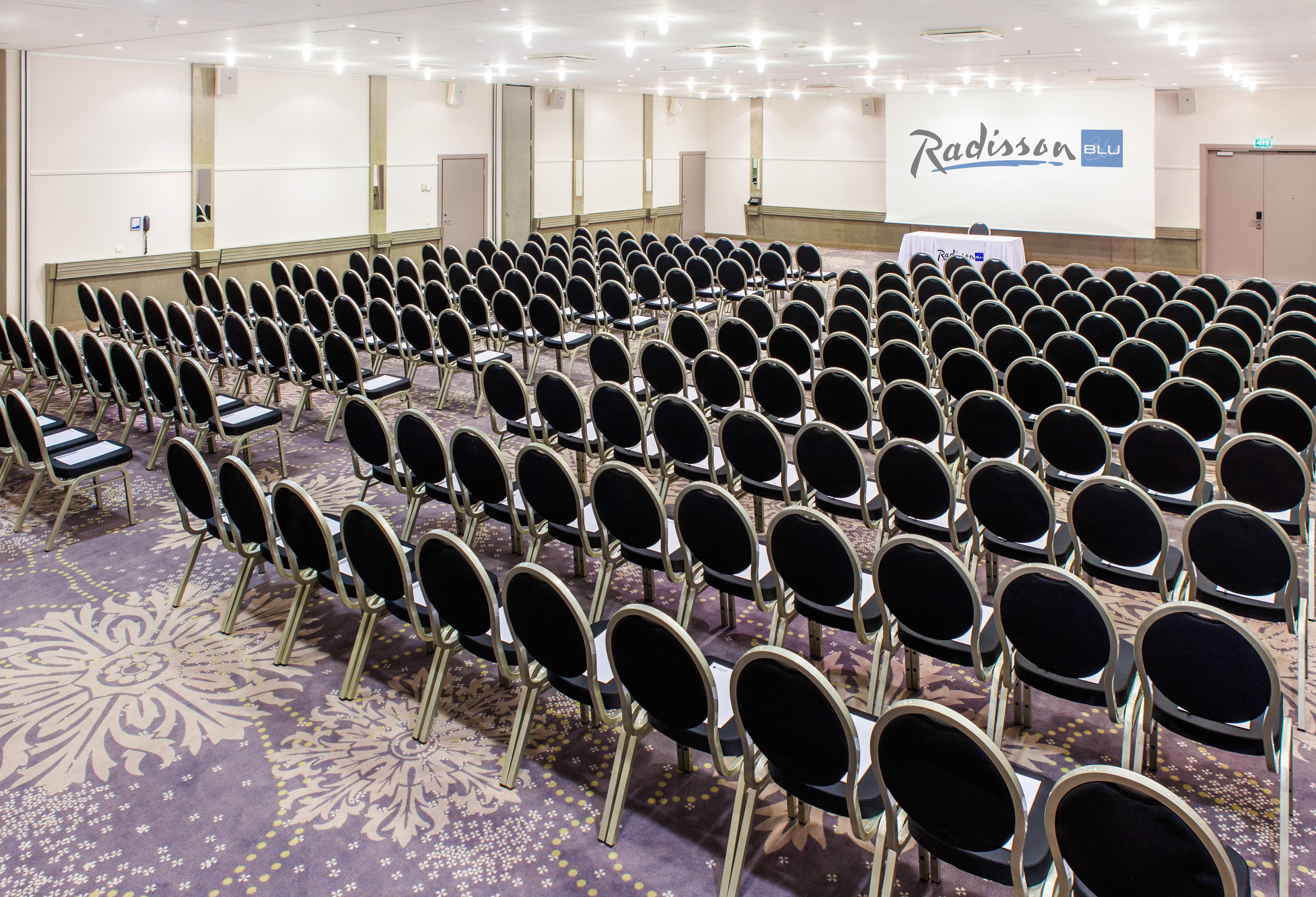 فندق راديسون بلو سكاي تالين المظهر الخارجي الصورة The photo shows a large conference room setup with rows of black chairs arranged facing a stage or presentation area at the front. The room has a light-colored carpet with a decorative pattern and is well-lit, featuring ceiling lights. In the backgro